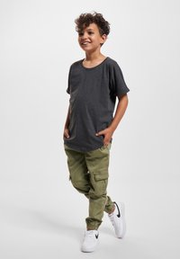 Dreng med krøllet hår, der smiler, hænderne i lommerne, iført en mørkegrå t-shirt, olivengrønne cargo-bukser og hvide sneakers med sort swoosh.