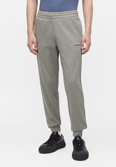 Мъж, носещ сиви спортни панталони EA7 Emporio Armani с еластични маншети и черни маратонки, стоящ на бял фон.
