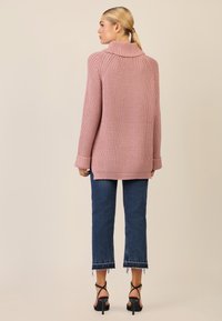 Übergroßer rosa gestrickter Pullover mit hohem Kragen, geripptem Muster und umgeschlagenen Bündchen, kombiniert mit hellblauen cropped Jeans und schwarzen Riemchensandalen.