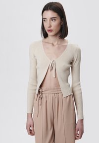 Beige gerippter Cardigan mit tiefem V-Ausschnitt und Bindebandverschluss vorne. Lange Ärmel und weicher, strukturierter Stoff. Kombiniert mit hellen Hosen.