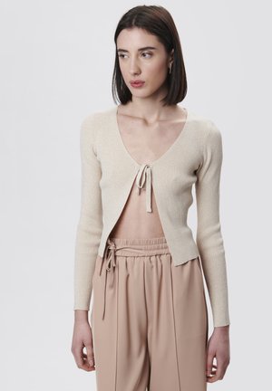 Beige gerippter Cardigan mit tiefem V-Ausschnitt und Bindebandverschluss vorne. Lange Ärmel und weicher, strukturierter Stoff. Kombiniert mit hellen Hosen.