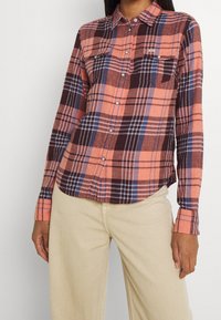 Chemise en flanelle à manches longues avec un motif à carreaux multicolores en orange, marron et bleu, dotée de boutons-pression et de deux poches poitrine.