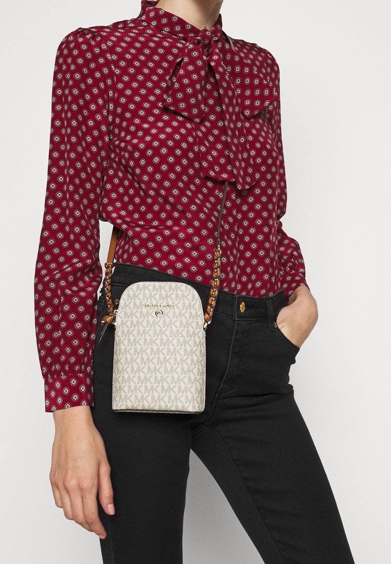Bolso bandolera beige con un patrón característico de MK, textura suave y una correa de cadena, combinado con una blusa burdeos con patrón y jeans negros.