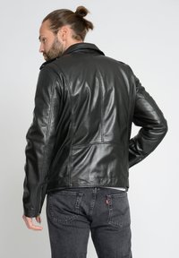 Czarna skórzana kurtka motocyklowa z prostym kołnierzem, zamkami na mankietach i szwami na plecach. Noszona na ciemnych dżinsach, pokazująca teksturowany materiał.