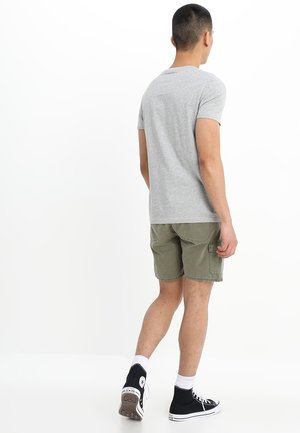 Maglietta grigia a maniche corte abbinata a pantaloncini verde oliva. L'outfit include scarpe da ginnastica nere alte e calzini bianchi alla caviglia.