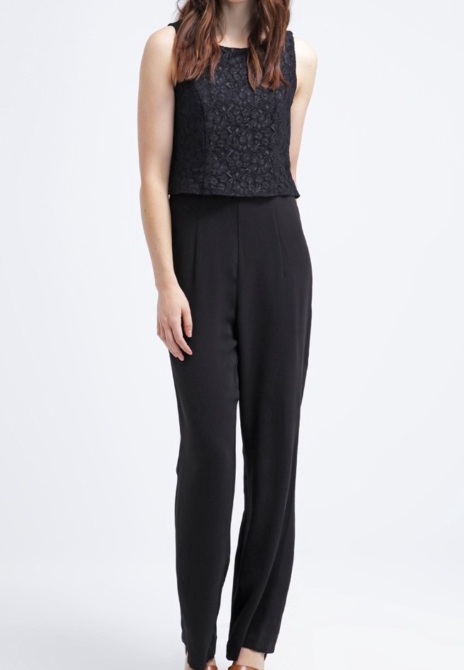 Femme portant un haut en dentelle noire sans manches et un pantalon large taille haute noir, debout sur un fond blanc uni.