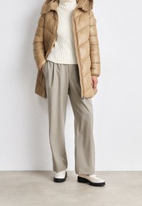Chaqueta acolchada beige con un acabado texturizado, suéter de punto crema clásico, pantalones de pierna ancha gris claro y mocasines blancos con suelas negras.