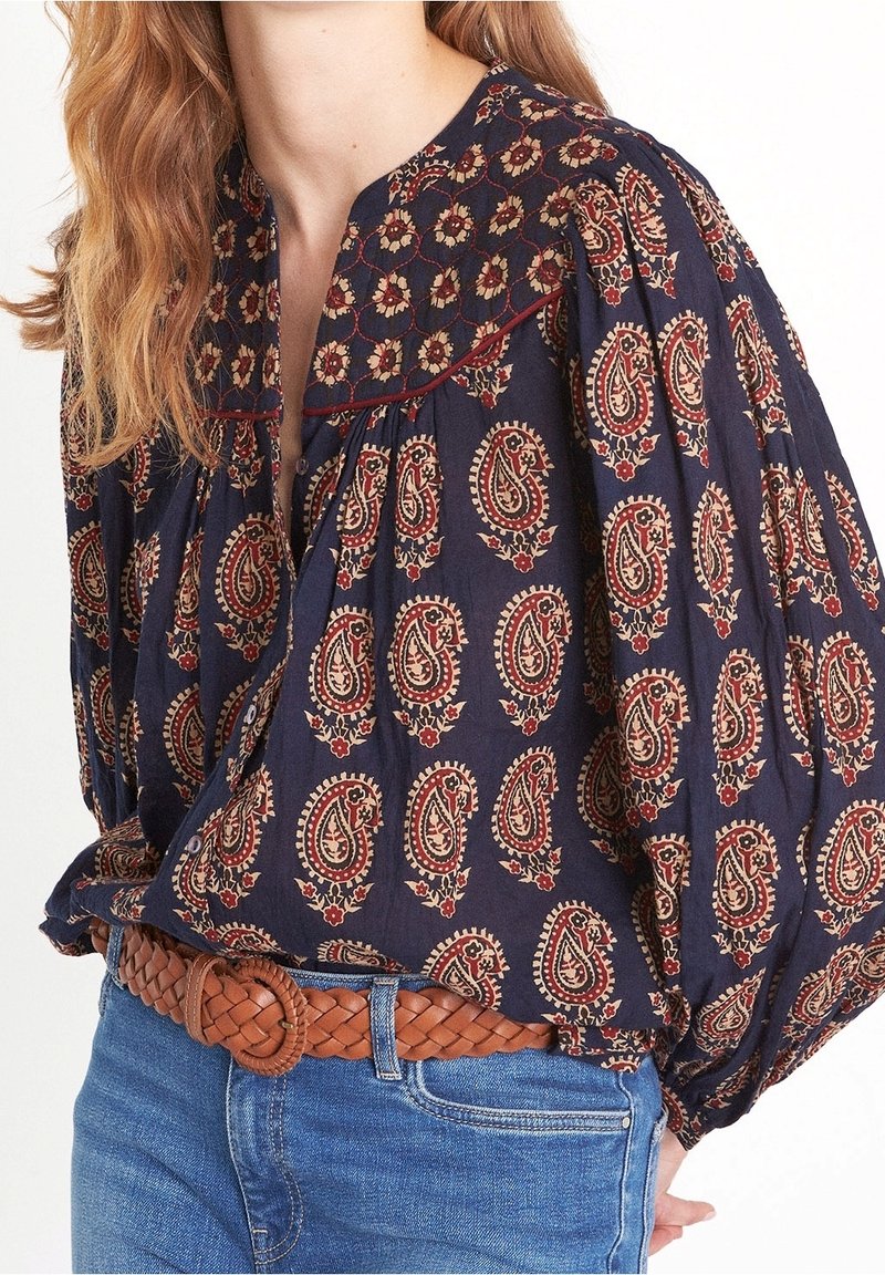 Blusa blu navy con stampa paisley in rosso e crema, presenta un colletto rotondo e maniche voluminose. Abbinata a una cintura in pelle intrecciata marrone.