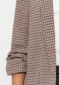 Marynarka z wzorem w pepitkę w kolorach różowym, czarnym i kremowym. Posiada podwinięte rękawy i gładką fakturę. Tkanina wydaje się sztywna i dopasowana.