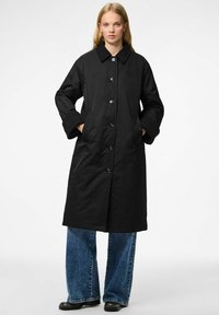 Cappotto lungo nero con colletto, bottoni frontali e tasche laterali. Tessuto liscio, vestibilità ampia, abbinato a jeans blu a gamba larga.