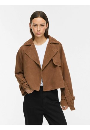 Jeune femme aux cheveux bruns raides porte une veste courte en daim marron, une chemise blanche et un pantalon noir taille haute sur un fond uni.