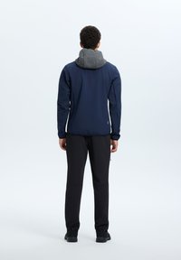 Giacca navy con cappuccio grigio, design slim fit, tessuto leggero e polsini elasticizzati. Abbinata a pantaloni neri e calzature scure.
