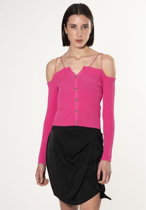 Top lungo a maniche con costine fucsia e spalle scoperte, scollatura a V e dettagli con bottoni sul davanti. Abbinato a una gonna nera a balze.