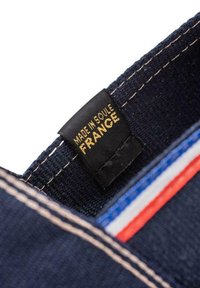 Tissu en denim foncé avec des coutures contrastées, arborant une étiquette noire indiquant "FABRIQUÉ À SOULE FRANCE" avec du texte doré et des bandes colorées en dessous.