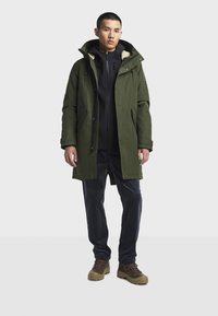 Giovane uomo che indossa un parka verde oliva con cappuccio foderato di pelliccia, giacca nera con zip, pantaloni scuri e scarponi da trekking marroni, in piedi su uno sfondo neutro.