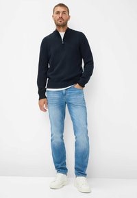 Maglione blu navy con zip, jeans azzurri e sneakers bianche. Il maglione presenta polsini e orlo a coste; i jeans sono slim-fit con una finitura sbiadita.