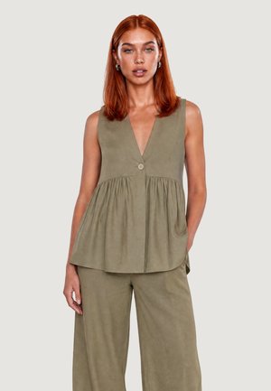 Donna con capelli rossi che indossa un top verde oliva senza maniche con bottoni e pantaloni a gamba larga abbinati, in piedi davanti a uno sfondo semplice.