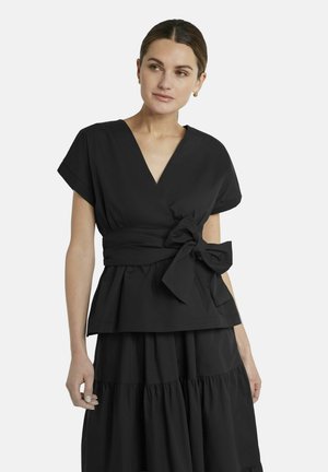 Frau trägt ein schwarzes Wickel-Top mit großem seitlichem Schleifenband und passendem, gestuftem schwarzem Rock, schaut leicht nach links.
