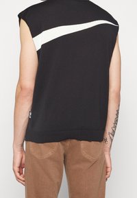 Haut en tricot sans manches noir avec une rayure diagonale blanche dans le dos, associé à un pantalon marron. Tissu texturé et coupe décontractée.