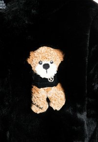 Tela de pelaje sintético negro con una aplicación de un oso de peluche. El oso tiene un cuerpo color marrón claro y un suéter negro, con rasgos faciales detallados.