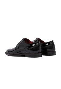 Een paar gepolijste zwarte leren schoenen met brogue-details en een rode binnenvoering, weergegeven vanaf de achterkant op een witte achtergrond.