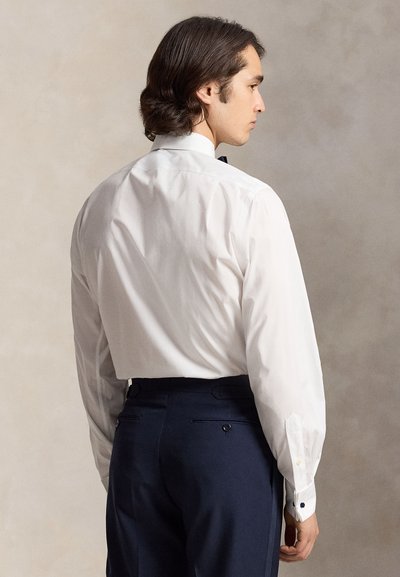 Uomo con capelli scuri che indossa una camicia bianca e pantaloni scuri, in piedi di profilo su uno sfondo uniforme.