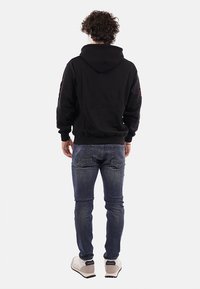 Felpa nera in cotone con tasca a canguro e polsini a coste. Indossata con jeans blu e sneakers chiare; mostrata da dietro.
