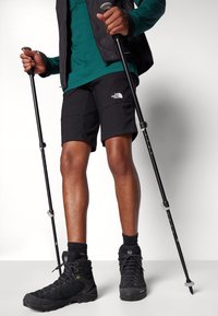 Shorts noirs, t-shirt à manches longues turquoise, bottes de randonnée noires et bâtons de trekking. Présente un logo sur les shorts, un design de chaussure texturé et des accents en mesh.