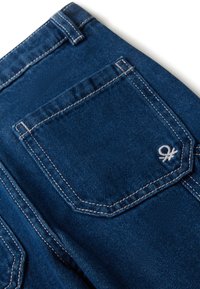 Modré džíny z denimu, zadní kapsa s bílým prošíváním a malým vyšitým bílým logem v dolním pravém rohu kapsy.