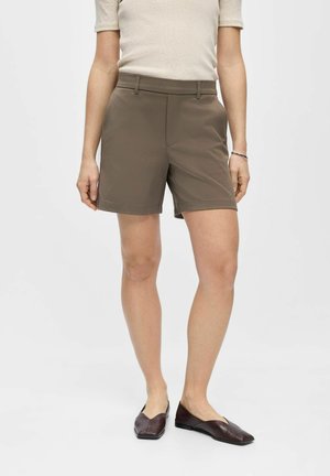Kvinde iført taupe skræddersyede shorts, beige kortærmet top og mørkebrune spidse flade sko mod en ensfarvet baggrund.