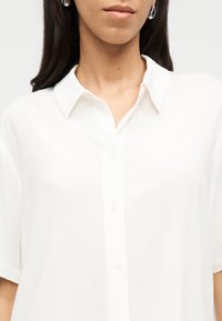 Γυναίκα που φοράει λευκό πουκάμισο με κοντά μανίκια και γιακά, ασημένια σκουλαρίκια κρίκους και μακριά σκοτεινά μαλλιά.