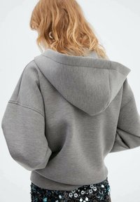 Sweat à capuche gris à coupe décontractée, en tissu doux, avec épaules tombantes et design simple. Détail de couture visible dans le dos.