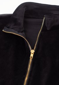 Zwarte fluwelen jas met een gedeeltelijk open gouden rits en een opgevouwen kraag op een witte achtergrond.