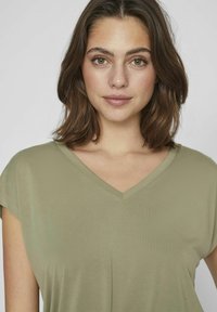 Ung kvinna med axellångt brunt hår iförd en olivgrön V-ringad topp, som tittar direkt in i kameran med ett neutralt uttryck.