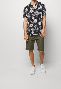 Mężczyzna w czarnej koszuli z krótkim rękawem w kwiaty, oliwkowych szortach i białych trampkach stoi na jednolitym tle.