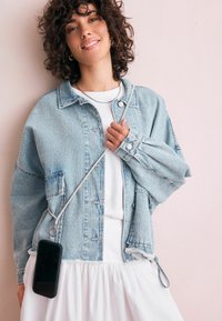 Krøllet hår kvinde smiler iført en lys denimjakke over en hvid kjole, mens hun holder en crossbody-telefon med sølv kædetras mod en lys pink væg.