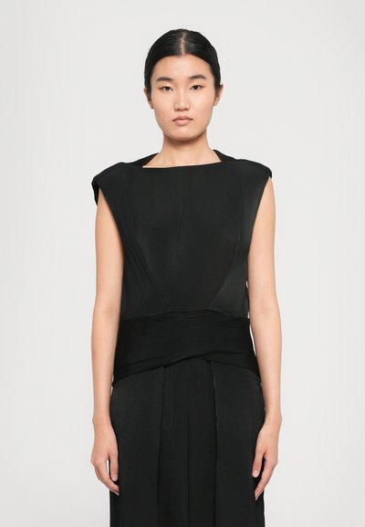 Femme aux cheveux noirs raides, portant une robe noire sans manches élégante avec une large ceinture, se tenant devant un fond gris uni.