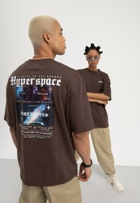 Un homme et une femme portant des t-shirts graphiques marron et des pantalons beiges se tiennent dans un studio ; l'homme montre l'impression au dos avec un design sur le thème de l'espace et un texte.