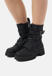 Botines negros de tobillo con suela gruesa, con cordones en la parte delantera, cremallera lateral y detalle de correa ajustable. Fabricados en un material suave y texturizado.