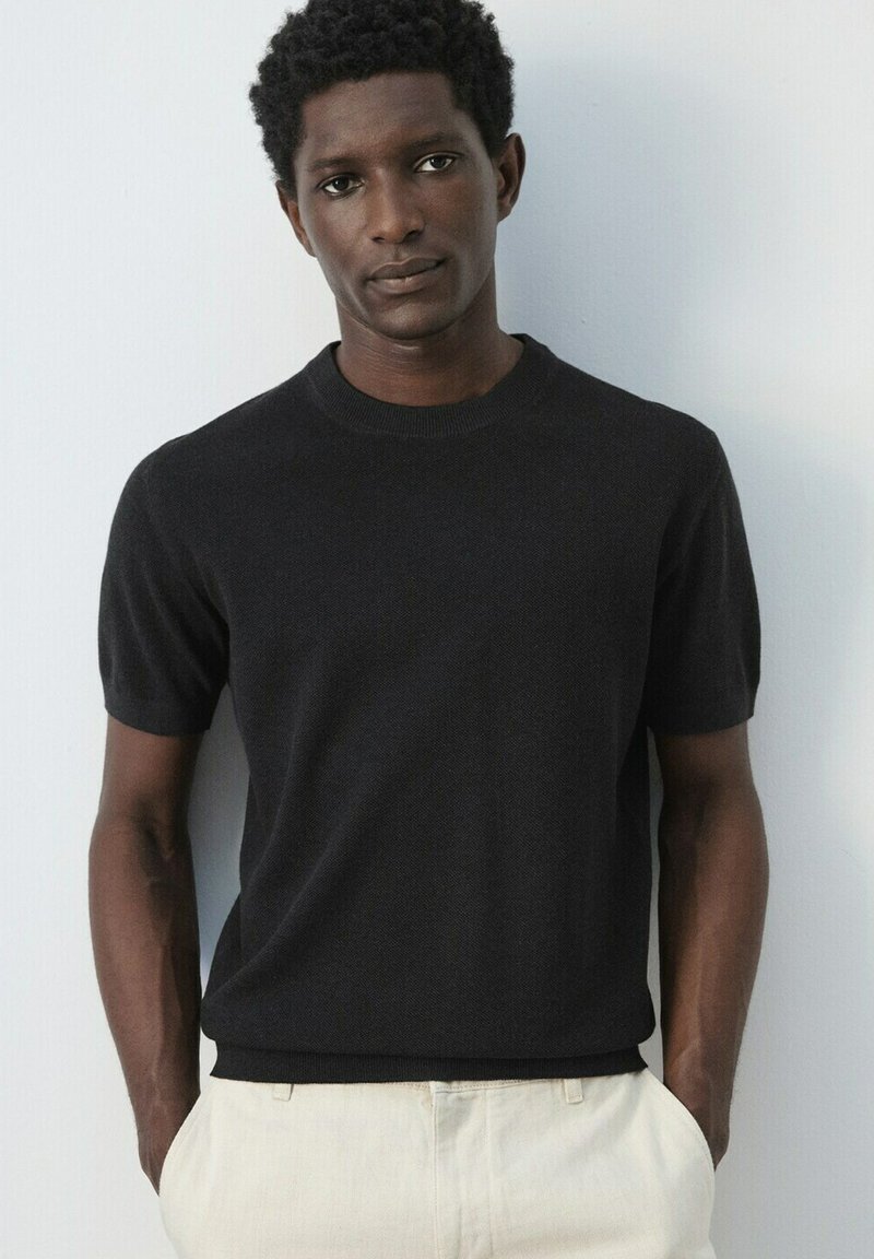 Homme aux cheveux courts et bouclés, portant une chemise noire à manches courtes et un pantalon de couleur claire, debout les mains dans les poches contre un mur blanc.