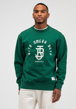 Mann trägt grünen Pullover mit weißem Text und Logo "Beta Omega Beta Butcher of Blue" und hellfarbige Hose, steht vor einfarbigem Hintergrund.
