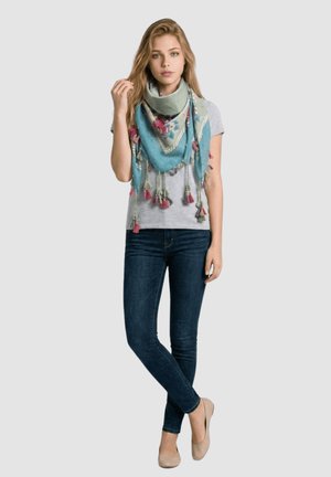 Mujer joven que lleva una camiseta gris claro, vaqueros pitillo oscuros, zapatos planos beige y una gran bufanda floral azul-verde con borlas.
