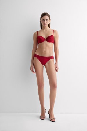 Femme debout contre un mur blanc, portant un soutien-gorge en dentelle rouge et une culotte assortie avec des sandales à talons noirs, faisant face.