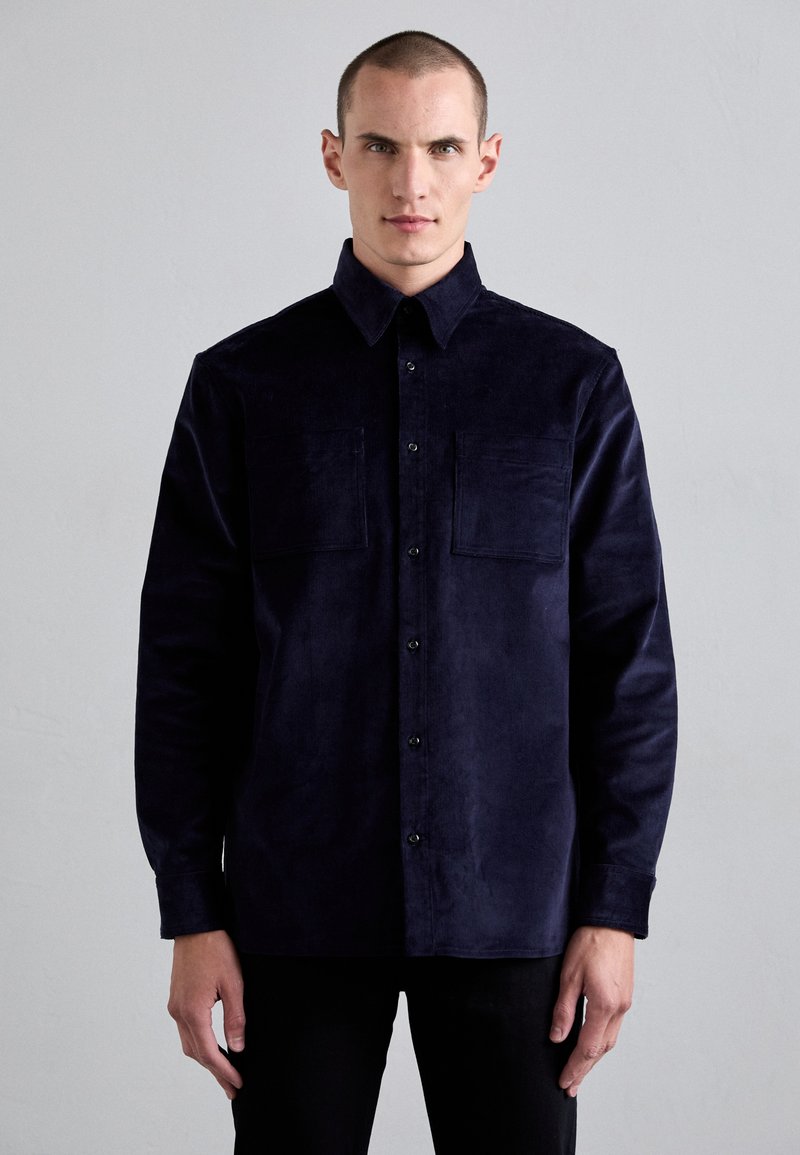 Camisa de pana de color azul marino, de manga larga, con botones en el frente y dos bolsillos en el pecho. Textura suave y cuello estructurado.