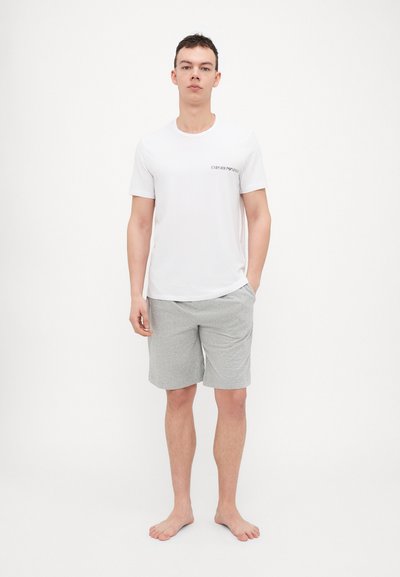 T-shirt bianco a maniche corte con la scritta "EMPORIO ARMANI" stampata in nero, abbinata a pantaloni corti in cotone grigi. Il modello è a piedi nudi.