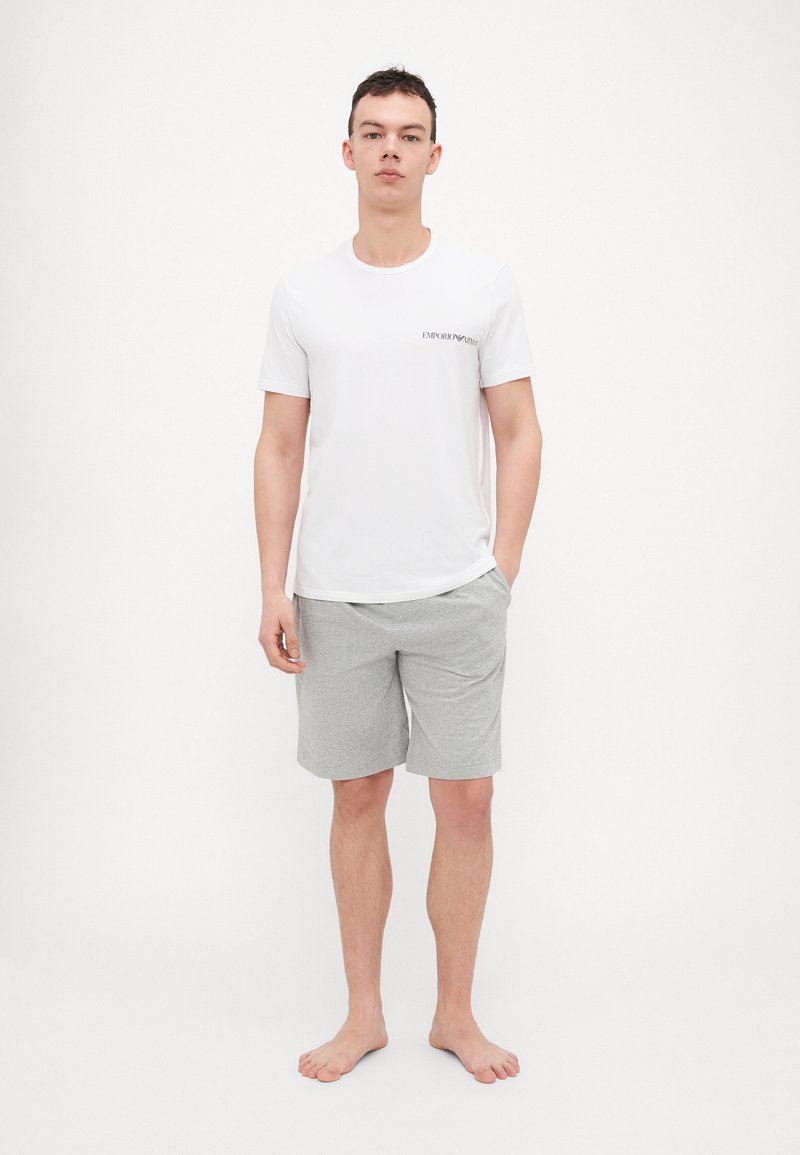 Weißes Kurzarm-T-Shirt mit dem schwarzen Aufdruck "EMPORIO ARMANI", kombiniert mit grauen Baumwollshorts. Das Modell steht barfuß.