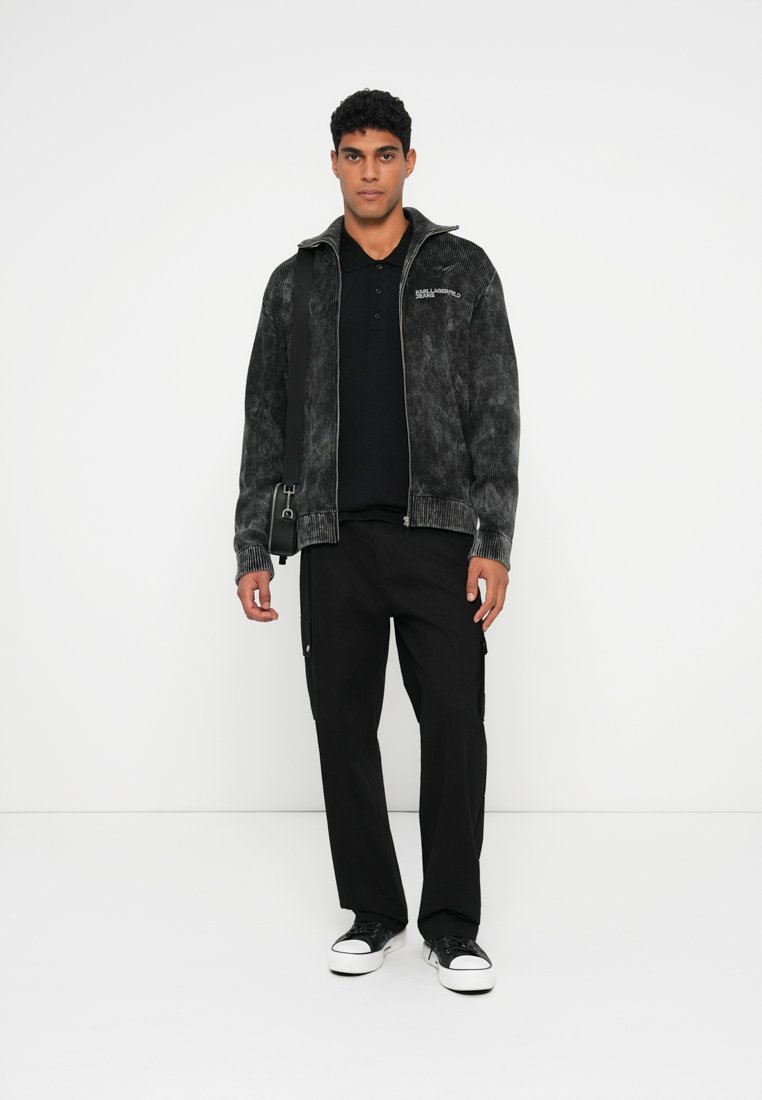 Veste noire texturée avec fermeture éclair à l'avant et col haut, portée sur un polo noir. Associée à un pantalon cargo noir et des baskets noires.