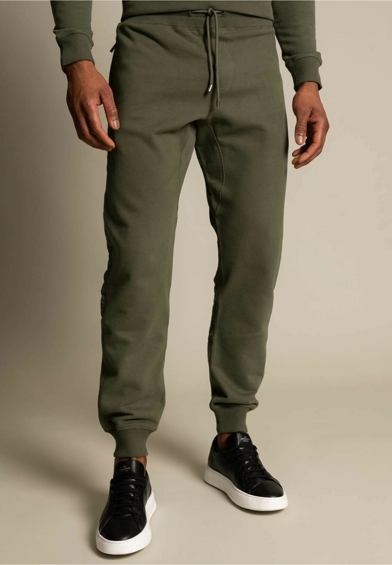 Olivegrüne Jogginghosen aus weichem Stoff mit schmal zulaufenden Beinen, elastischen Bündchen und verstellbarem Kordelzug am Bund. Kombiniert mit schwarzen Turnschuhen.