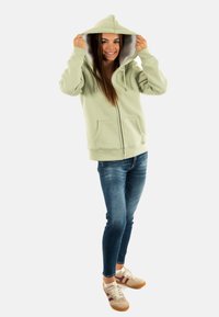 Un sweat à capuche zippé vert clair avec un intérieur doux, une capuche avec cordon de serrage et des poches avant. Porté avec un jean foncé et des baskets beiges.
