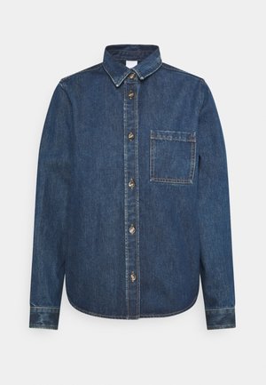 Chemise en denim bleu à manches longues avec boutonnière devant et poche poitrine sur fond clair uni.